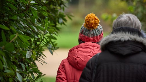 Head and shoulders of two people from behind, walking through a garden, past an evergreen hedge. The day is dull, they're wearing thick winter coats, one has a colourful woollen bobble hat on.